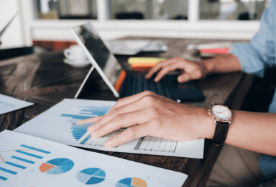 The image shows a man's hands typing on his computer and looking at a bunch of data visualization reports laid out on his desk in front of him.
